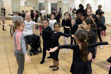Professeur de danse en fauteuil roulant animant un cours de jeunes filles, parents en fond