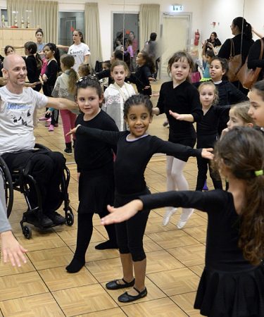 Professeur de danse en fauteuil roulant animant un cours de jeunes filles, parents en fond