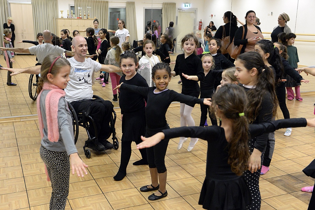 Professeur de danse en fauteuil roulant animant un cours de jeunes filles, parents en fond